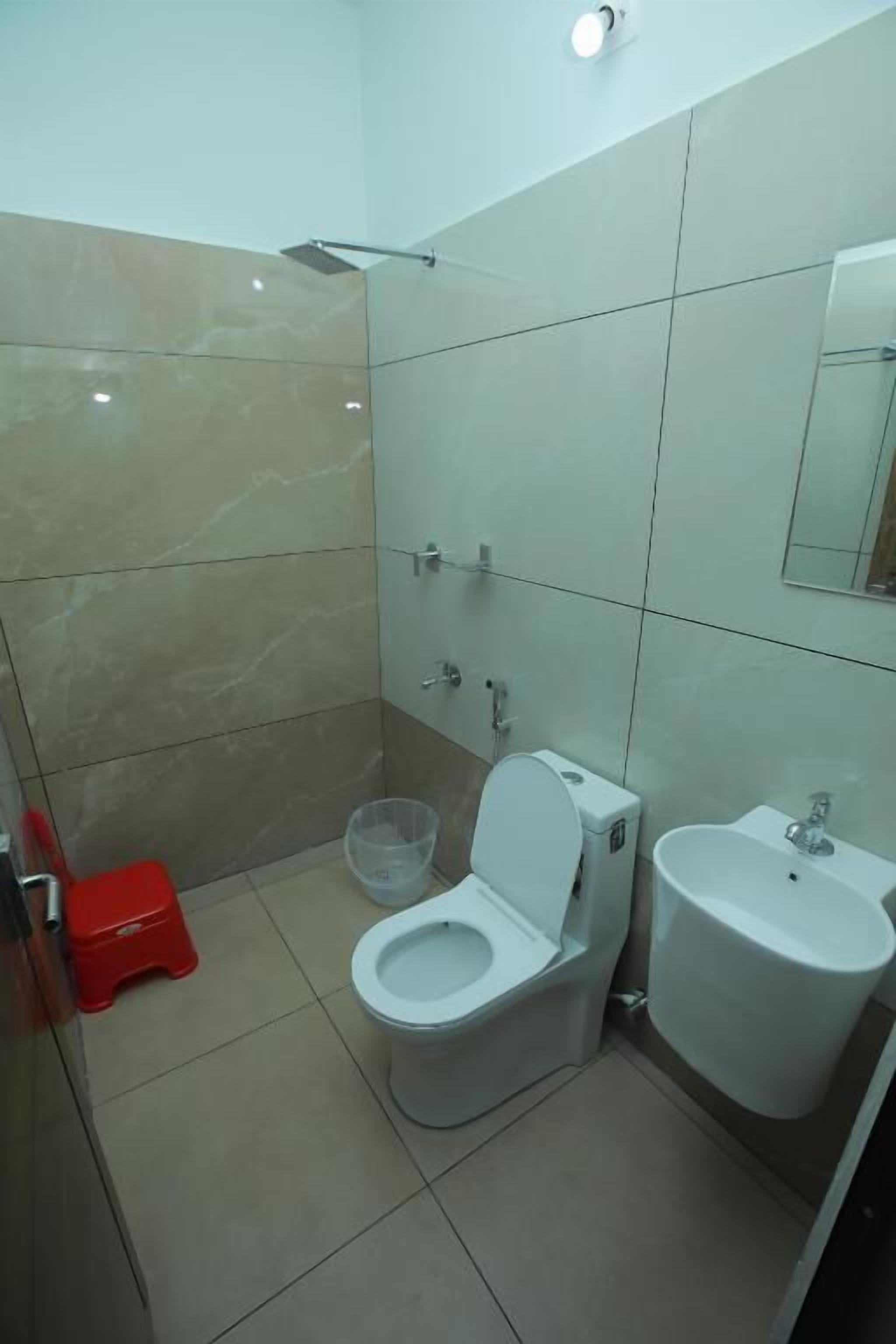 Casa de banho com sanita, lavatório, chuveiro e um banquinho vermelho.