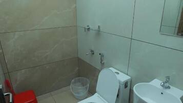 Casa de banho com sanita, lavatório, chuveiro e um banquinho vermelho.