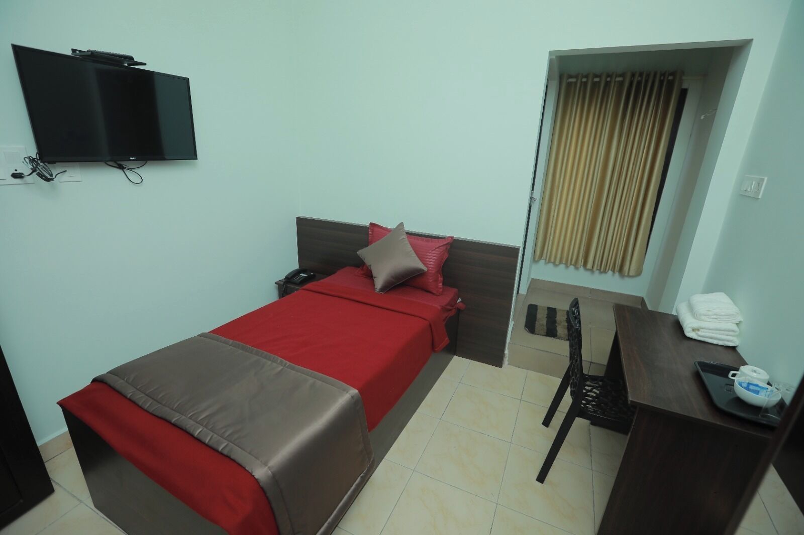 Una cama individual con colcha roja y marrón, un escritorio con una silla, un televisor montado en la pared y una ventana con cortinas.