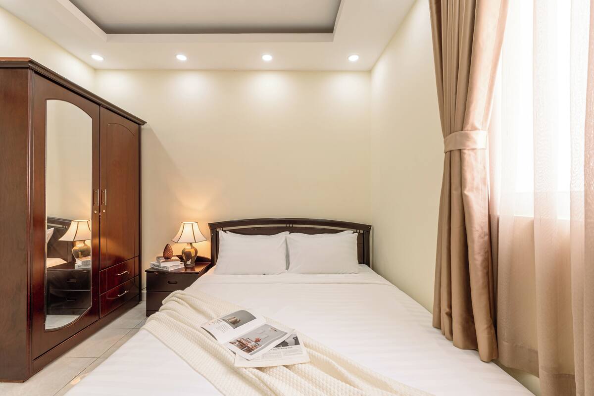 A bedroom with a bed, a wardrobe, a nightstand with a lamp, and a window with curtains.