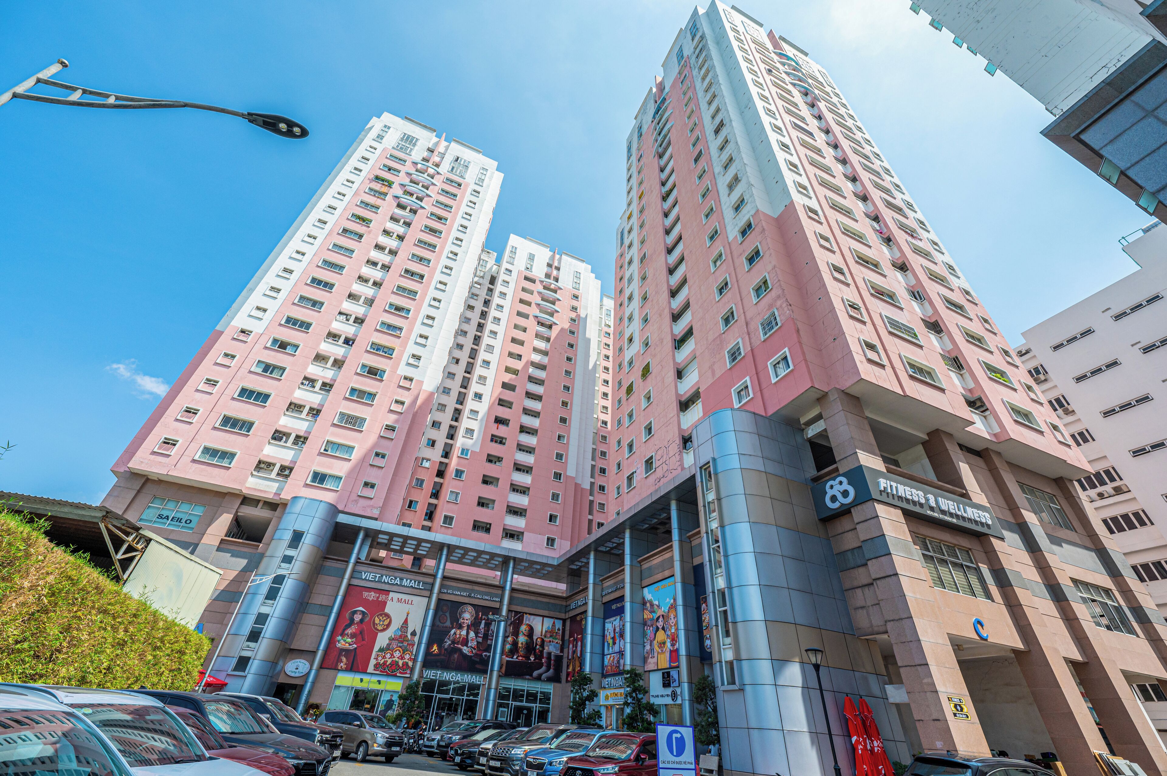 Un immeuble à plusieurs étages, avec une grande enseigne indiquant la présence d’un centre de remise en forme, une vaste publicité pour le centre commercial Viet Nam Mall, et plusieurs voitures garées devant.