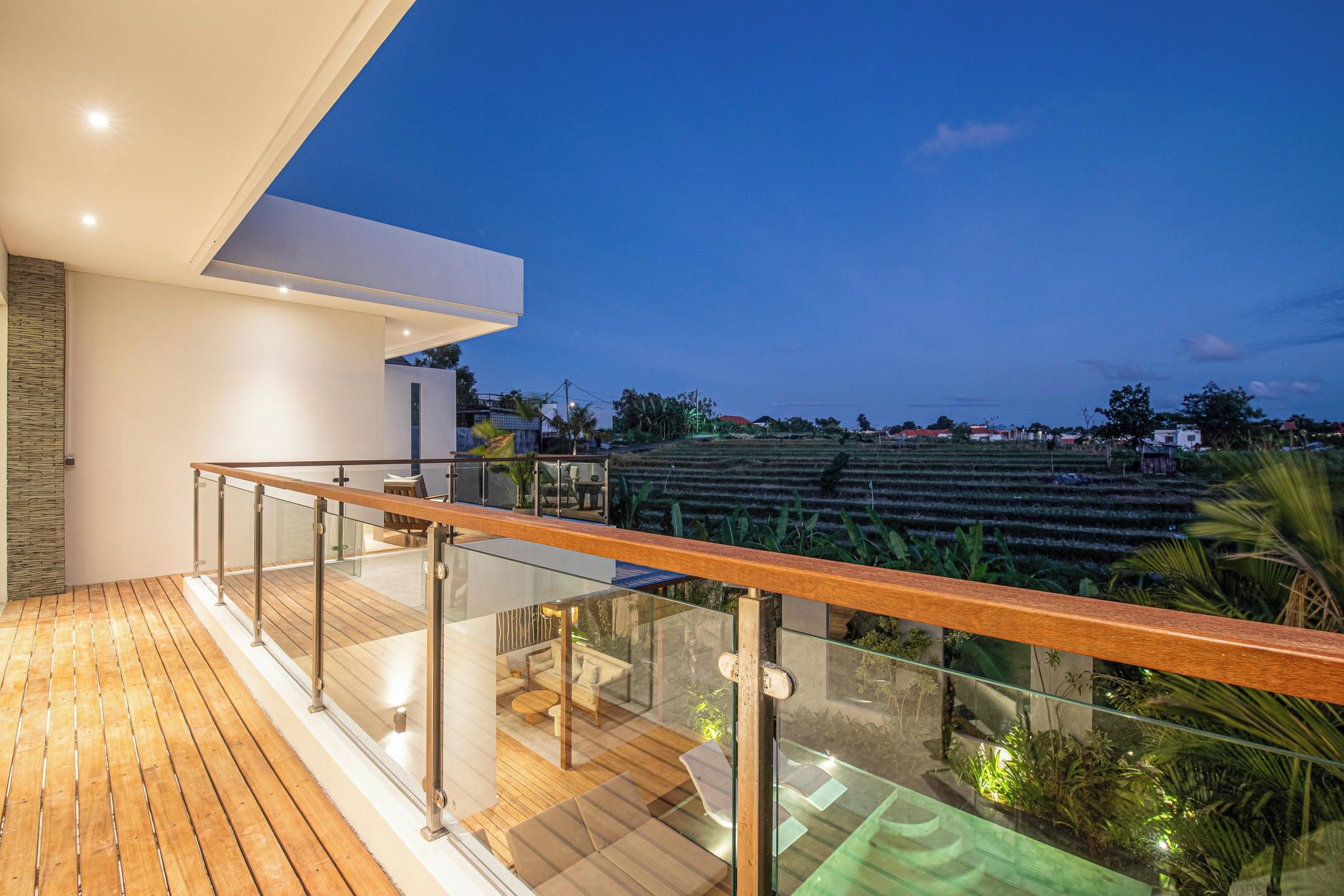 Un balcon moderne avec des garde-corps en verre, un plancher en bois et une vue sur des champs en terrasses.