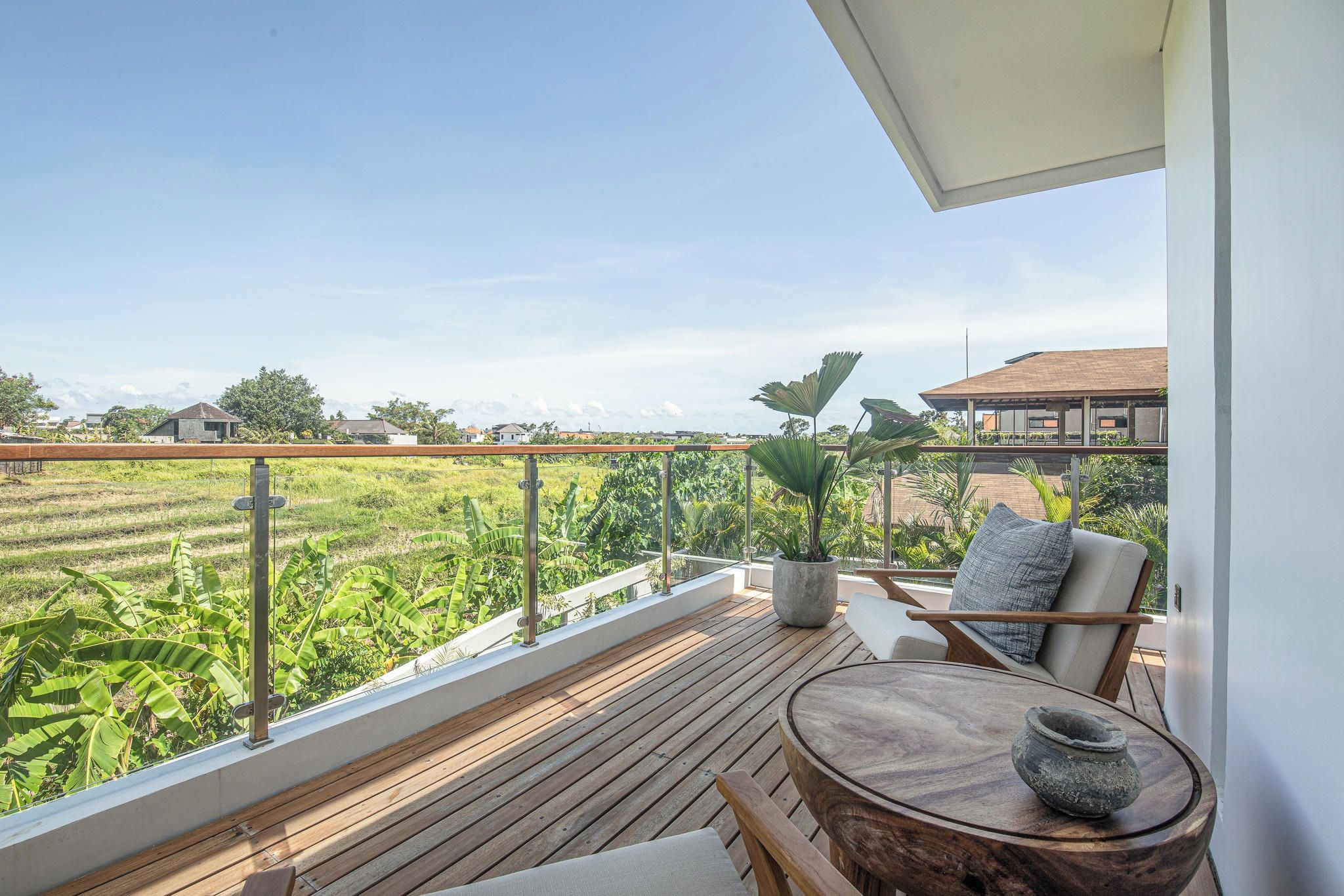A balcony with a view of a green field, a wooden table, a chair, and a potted plant.