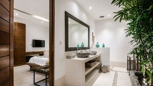Une salle de bain moderne avec un grand miroir, un lavabo agrémenté de vases décoratifs, un téléviseur en arrière-plan et une plante sur la droite.