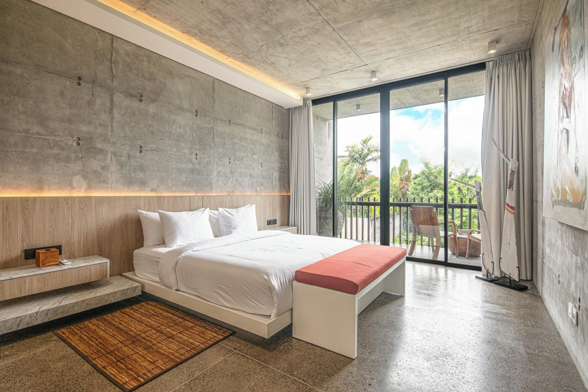A modern bedroom with a large bed, a bench, a nightstand, and a view of the outdoors through a sliding glass door.