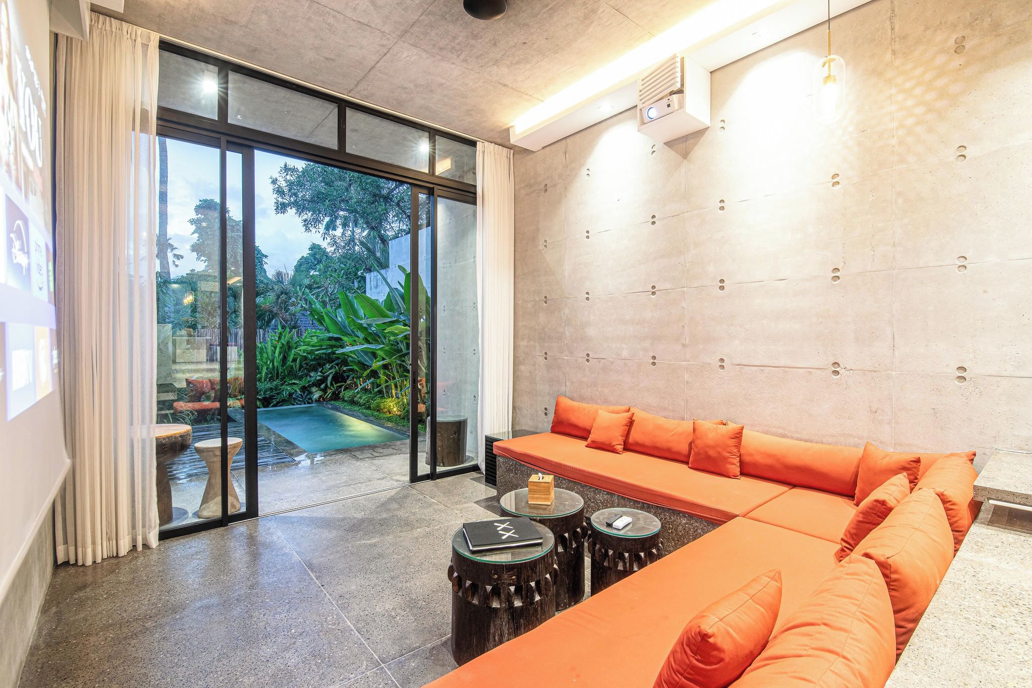 Una sala de estar moderna con un sofá grande color naranja, un ventanal de vidrio que da a una piscina y una pared de concreto con un accesorio de luz.