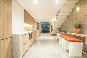 A modern kitchen with wooden cabinets, a marble countertop, and a central island. There are white stools with orange cushions and a large vase with flowers on the right side. - Modern 2BR Villa Enclosed Living, Heart of Canggu (Kerobokan)