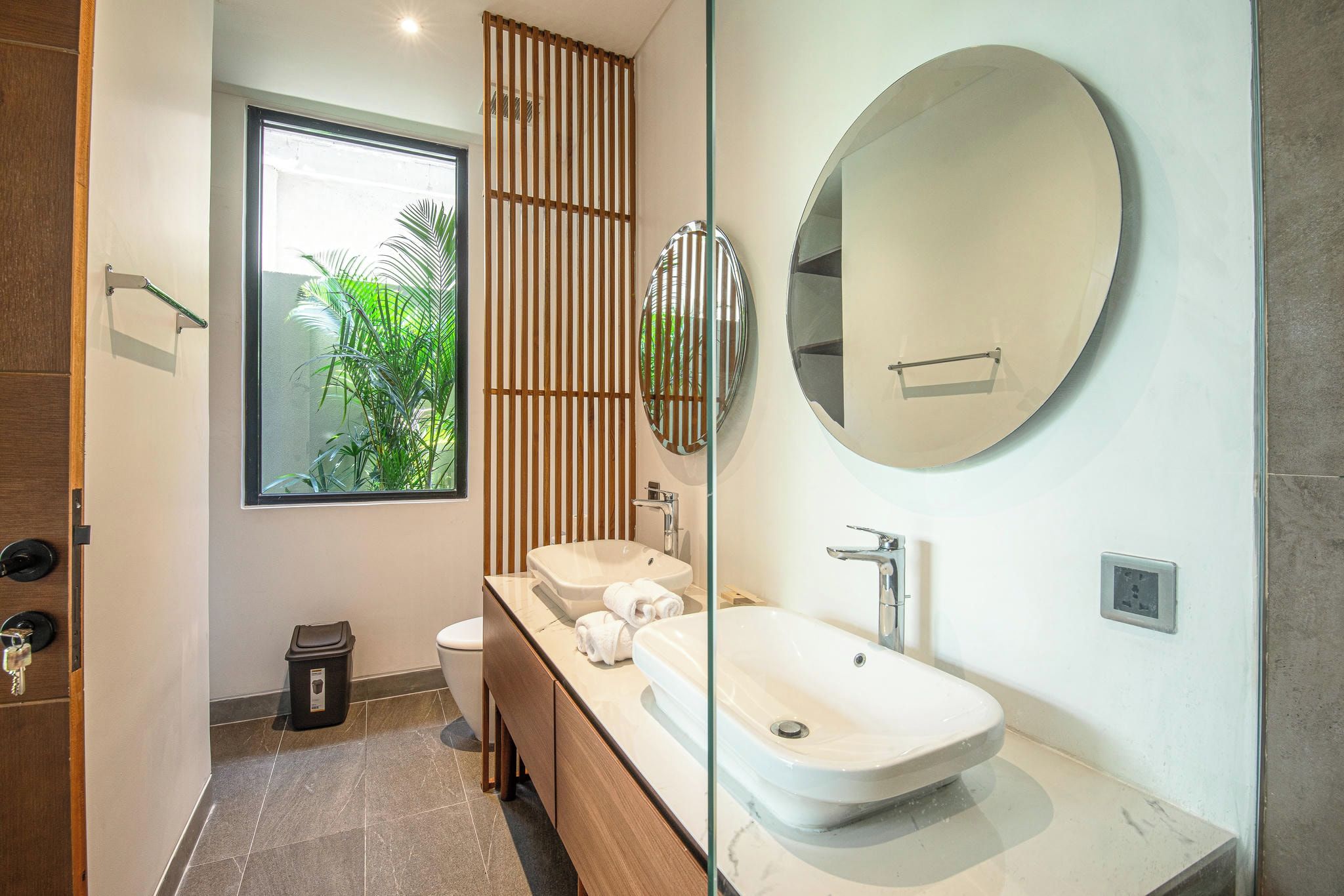 Une salle de bain moderne avec une grande fenêtre, un miroir rond, un lavabo et des toilettes.