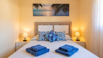 Een opgemaakt bed met blauwe handdoeken, gestreepte kussens en een schilderij van een zonsondergang erboven.