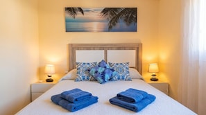 Un lit bien fait, avec des serviettes bleues, des coussins à motifs et un tableau représentant un coucher de soleil au-dessus.