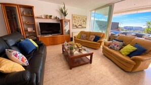 Uma sala de estar com um sofá de couro preto, um sofá de couro marrom e uma mesa de centro de vidro. Há uma televisão em um móvel de madeira, e um quadro na parede.