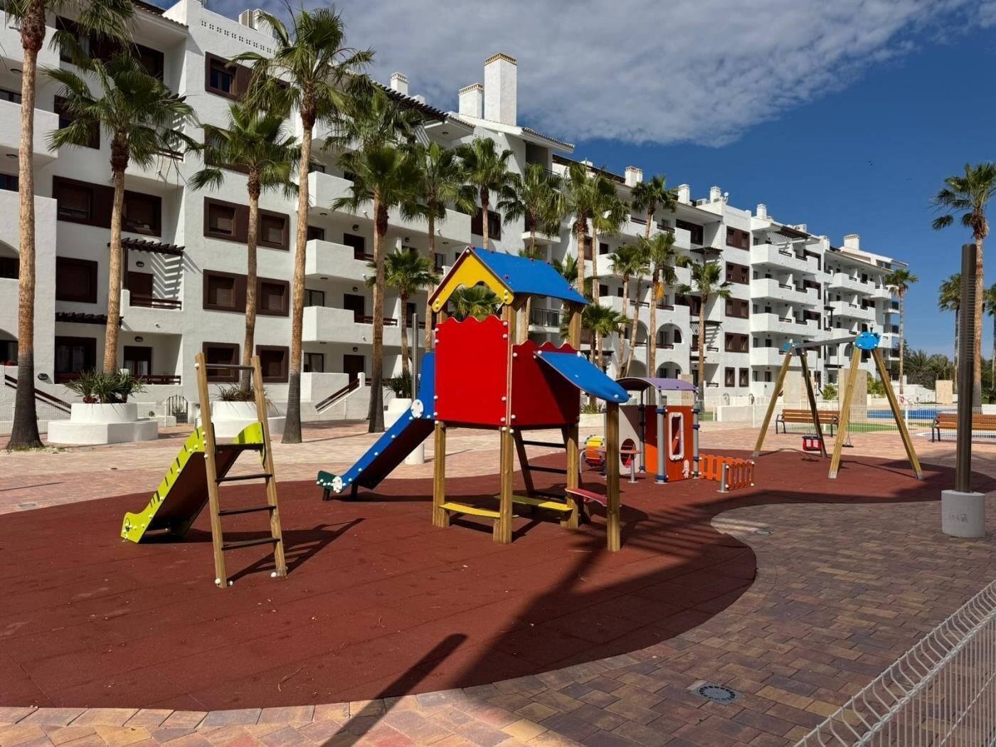 Un parque infantil con tobogán y estructuras para escalar, rodeado de palmeras y un edificio de varios pisos con balcones.