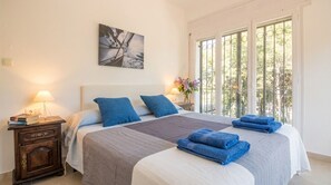 A hotel room with a large bed, a nightstand with a lamp, a painting on the wall, and a window with a view of trees. - Villa Playa del Sur · 250m from the sea · 7 Suites · +19 guests · Views 10 P (Capafonts)