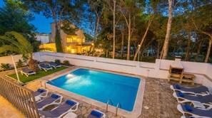 Piscina com espreguiçadeiras e deck de pedra, cercada por árvores e um edifício ao fundo.