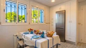 A kitchen with a stainless steel refrigerator, white cabinets, a dining table with a blue tablecloth, and a window with a view of trees. - Villa Playa del Sur · 250m from the sea · 7 Suites · +19 guests  · Views 14 P (Capafonts)
