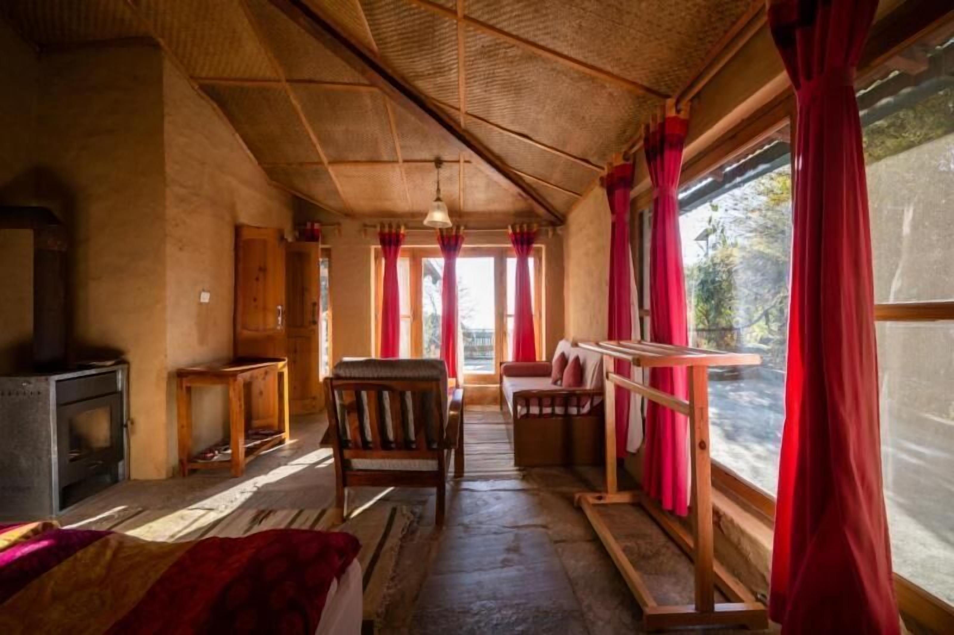 A rustic cabin interior with a wooden ceiling, a fireplace, a bench, and a window with red curtains.