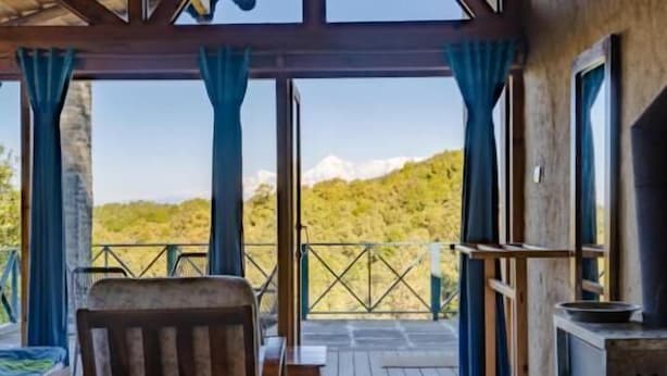 A wooden deck with a chair and a table, a fireplace, and a view of a mountainous landscape. - Binsar Forest Retreat (Binsar)