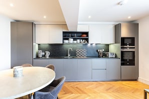 Eine moderne Küche mit grauen Schränken, einer schwarzen Arbeitsplatte, einem eingebauten Backofen und einem Essbereich mit einem runden Tisch und Stühlen.