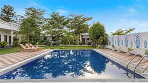 A rectangular swimming pool with a checkered deck, surrounded by trees and a white building with blue decorative patterns. - Alaya stays Haven Villa 3BHK with Pool (Bhankrota)