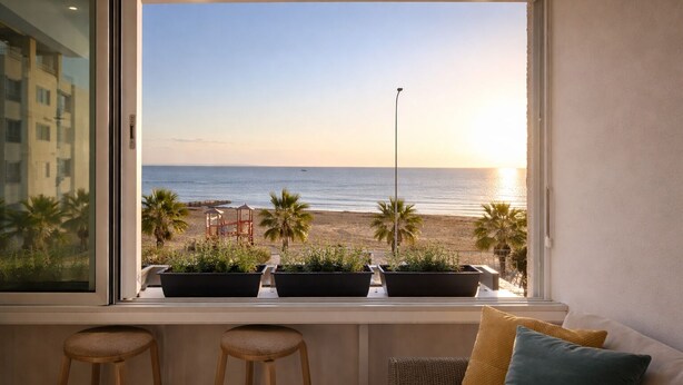 Ein Zimmer mit Blick auf den Strand, ein Fenster mit Blumentöpfen und ein Sofa mit Kissen.