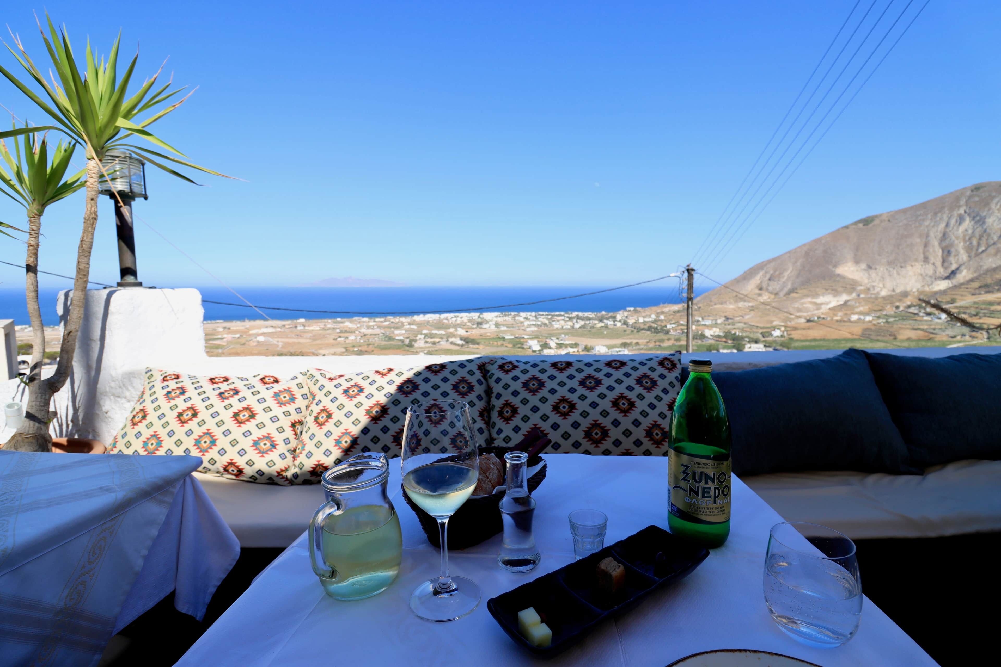 Ett bord med en flaska ZUNONE NEPO, ett glas vitt vin, ett glas vatten och en desserttallrik, placerat med en vacker utsikt över havet och kullarna i bakgrunden.