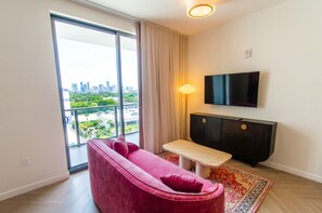 Ein modernes Wohnzimmer mit einer Couch, einem Fernseher, einem kleinen Tisch und Blick auf eine Stadtlandschaft durch ein großes Fenster.