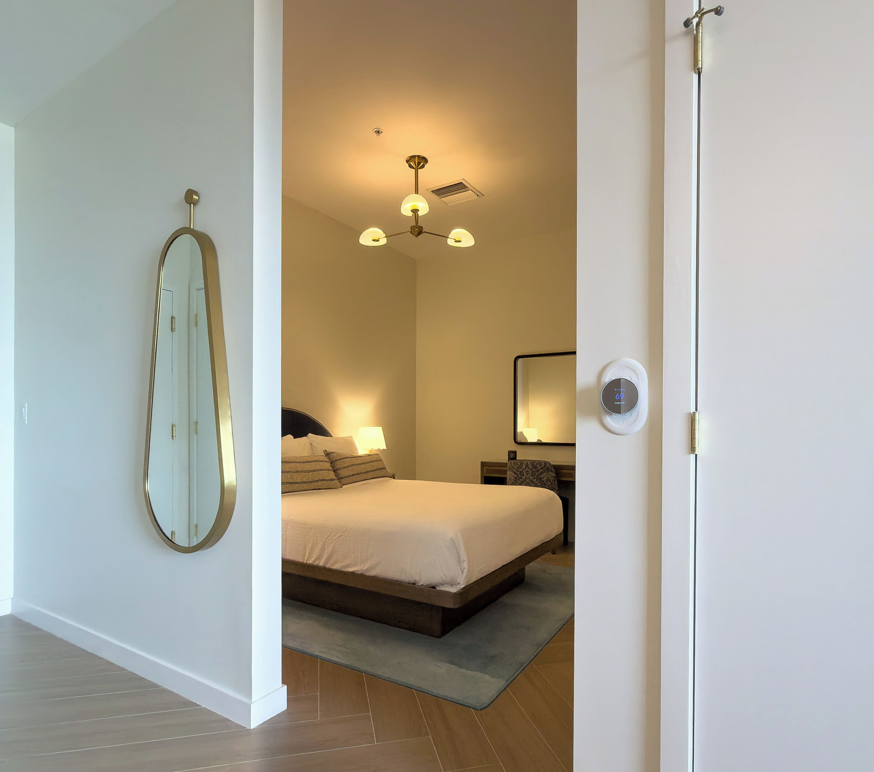A bedroom with a bed, bedside table, lamp, mirror, ceiling light, and a thermostat.