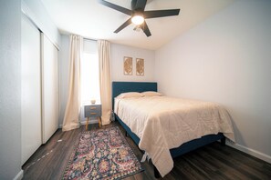 A bedroom with a bed, a ceiling fan, a nightstand, a window with curtains, and a patterned rug.
