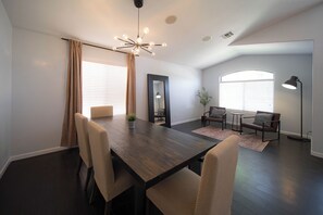 A modern dining area with a dark wooden table, beige chairs, a chandelier, and large windows with blinds.