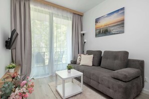 Ett modernt vardagsrum med en grå soffa, ett vitt sidobord, en krukväxt och en tavla som föreställer en solnedgång.