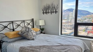 Une chambre à coucher avec un lit, des oreillers, une lampe de chevet, une œuvre d’art murale et une fenêtre donnant sur la ville.