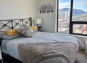 Un dormitorio con cama, almohadas, una lámpara de noche, una obra de arte en la pared y una ventana con vista a la ciudad.