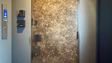 Una puerta con superficie texturizada y el número 901. Hay una pantalla digital en la pared a la izquierda, y un pequeño dispositivo montado en la pared sobre la puerta.