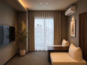 Een moderne hotelkamer met een bank, een bloempot, een flatscreen televisie en een groot raam met gordijnen.