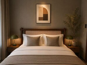 Una cama bien hecha con cabecera, dos mesitas de noche con lámparas, un cuadro enmarcado en la pared y una planta en maceta.
