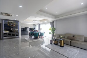 Ein modernes Wohnzimmer mit einer Couch, einem Essbereich mit Stühlen, einer Küche im Hintergrund und einem großen Fenster mit Vorhängen.