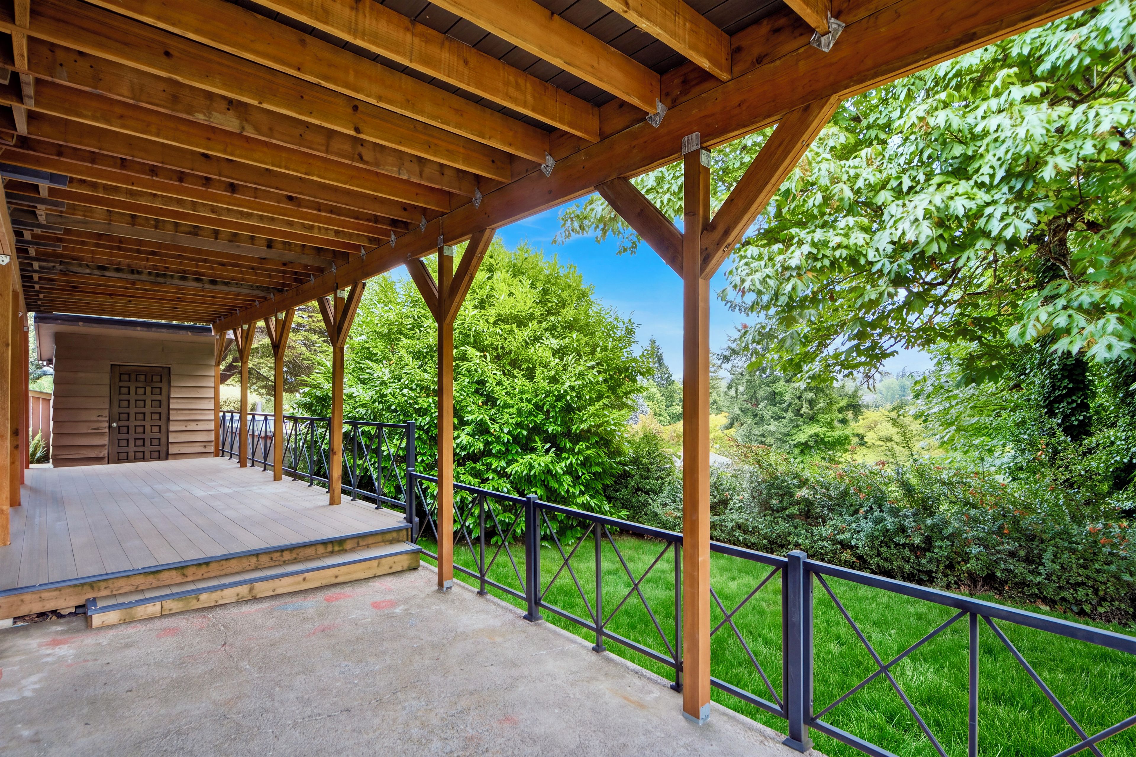A covered outdoor area with a wooden deck, metal railing, and a view of lush greenery.