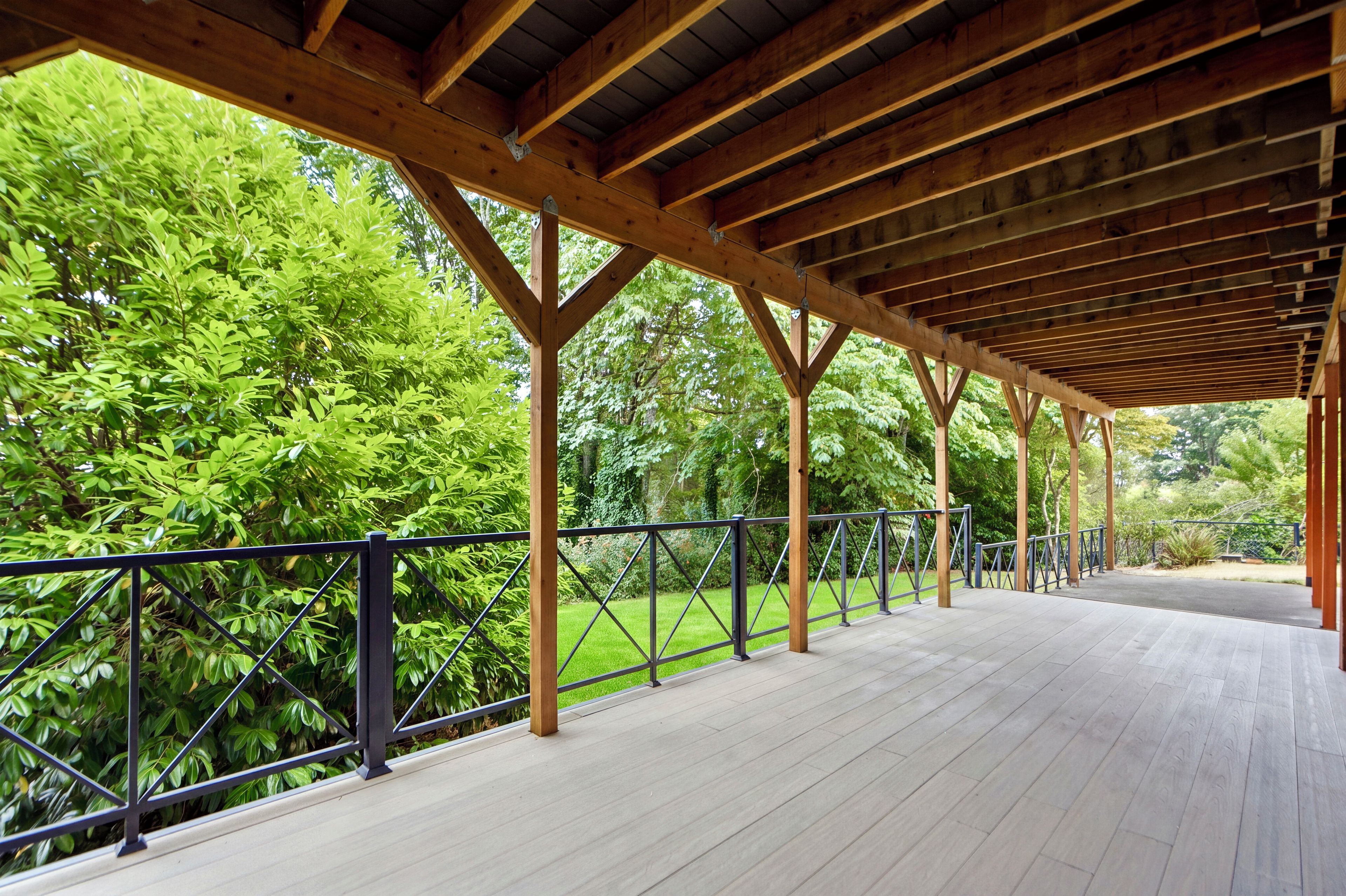 A covered outdoor area with a wooden deck, metal railing, and lush greenery in the background.