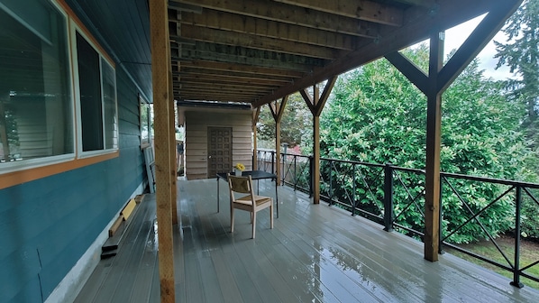 A covered outdoor deck with a table and chairs, wooden beams, and a view of greenery.
