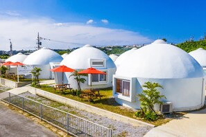 Fileira de edifícios em forma de cúpula, com mesas de piquenique e guarda-sóis vermelhos, cercada por vegetação.