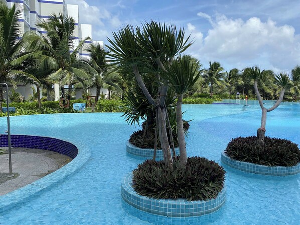 A swimming pool with palm trees and a tiled edge, surrounded by greenery and a building in the background. - Arena Cam Ranh Beachfront Condotel (Bac Cam Ranh)