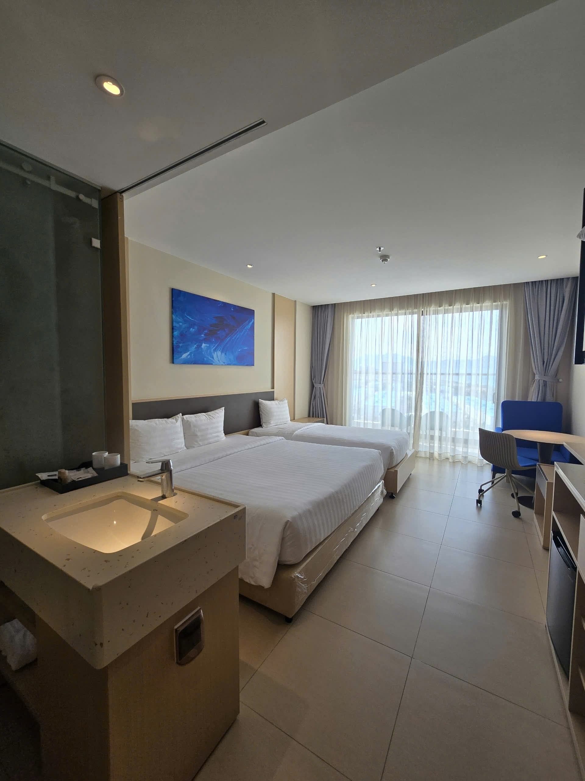 Quarto de hotel com uma cama grande, uma escrivaninha, uma cadeira, banheiro com pia e uma janela ampla com cortinas.