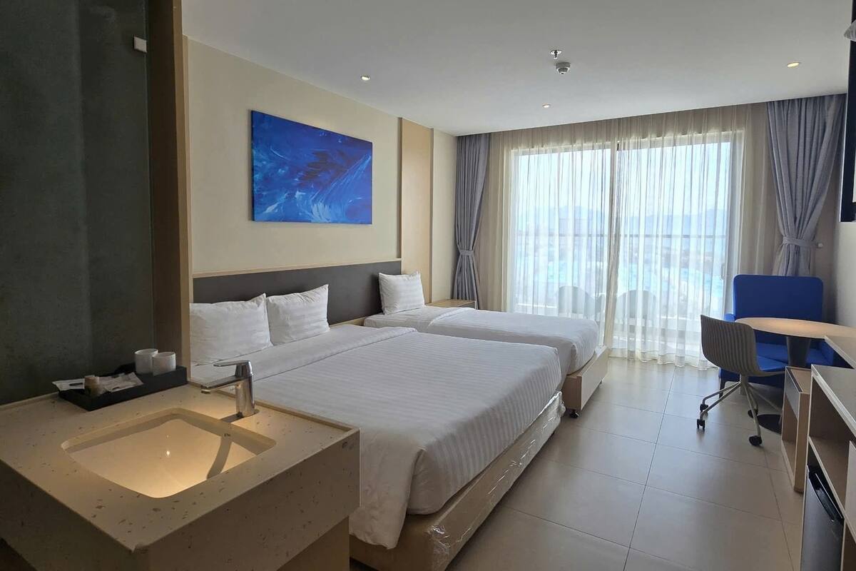 Habitación de hotel con una cama grande, un escritorio, una silla, un baño con lavamanos y un ventanal con cortinas.