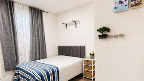 Ein Schlafzimmer mit einem Bett, einem Nachttisch mit Pflanze, einem Bücherregal und einem Fenster mit Vorhängen.