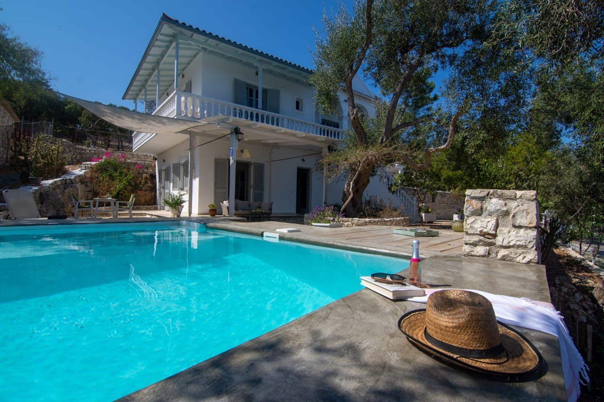 A swimming pool with a white house in the background, a stone wall, and a hat on the poolside.