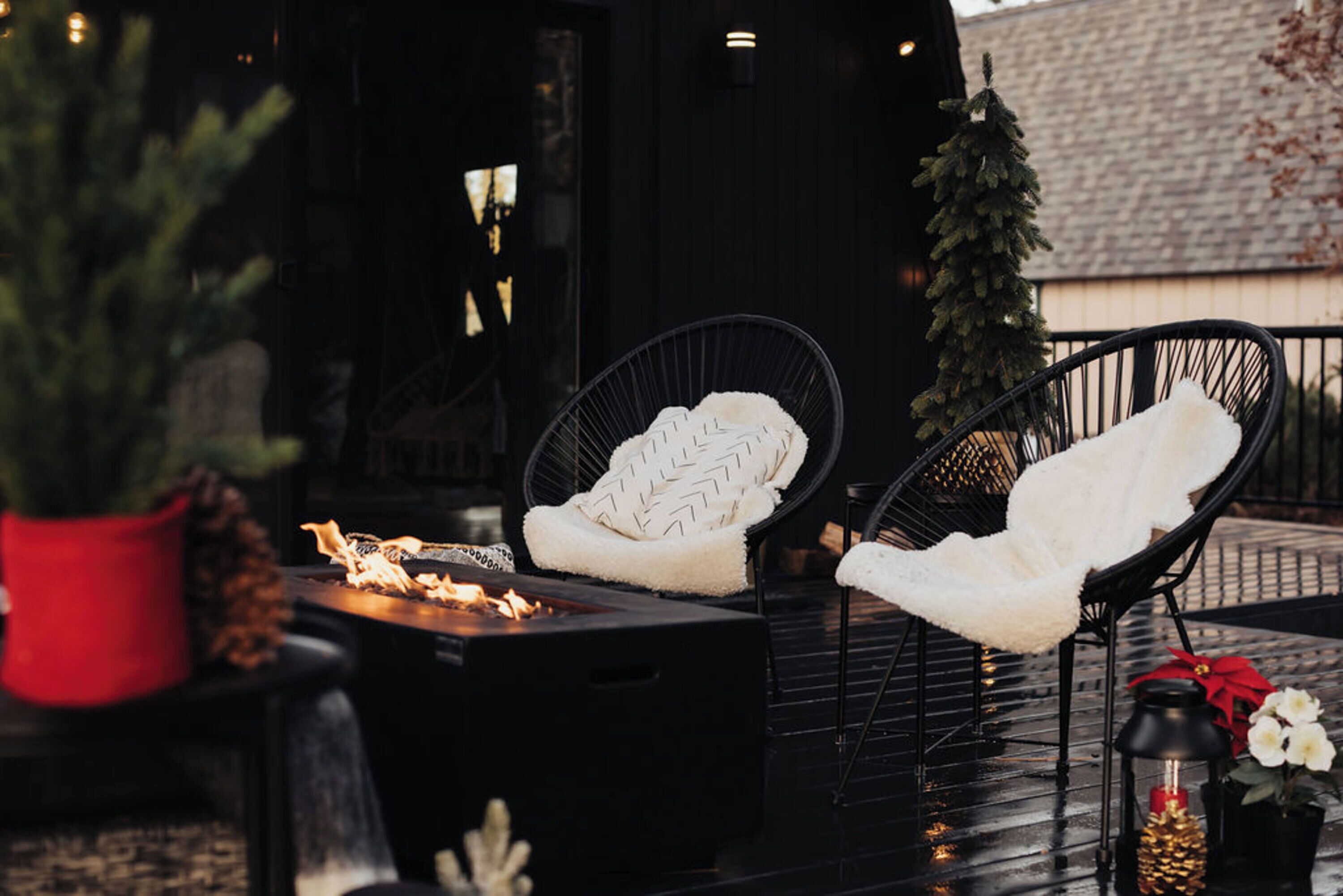 A cozy outdoor seating area with a fire feature, two chairs with white cushions, a small Christmas tree, and decorative lanterns.