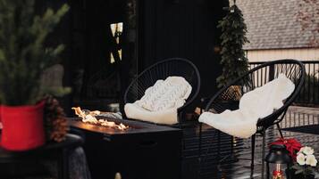 A cozy outdoor seating area with a fire feature, two chairs with white cushions, a small Christmas tree, and decorative lanterns.