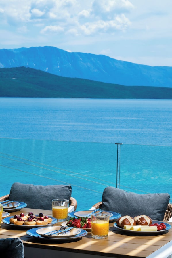 A dining table with plates of food, a glass of orange juice, and a view of a lake with mountains in the background. - Blue Horizon (Lefkada)