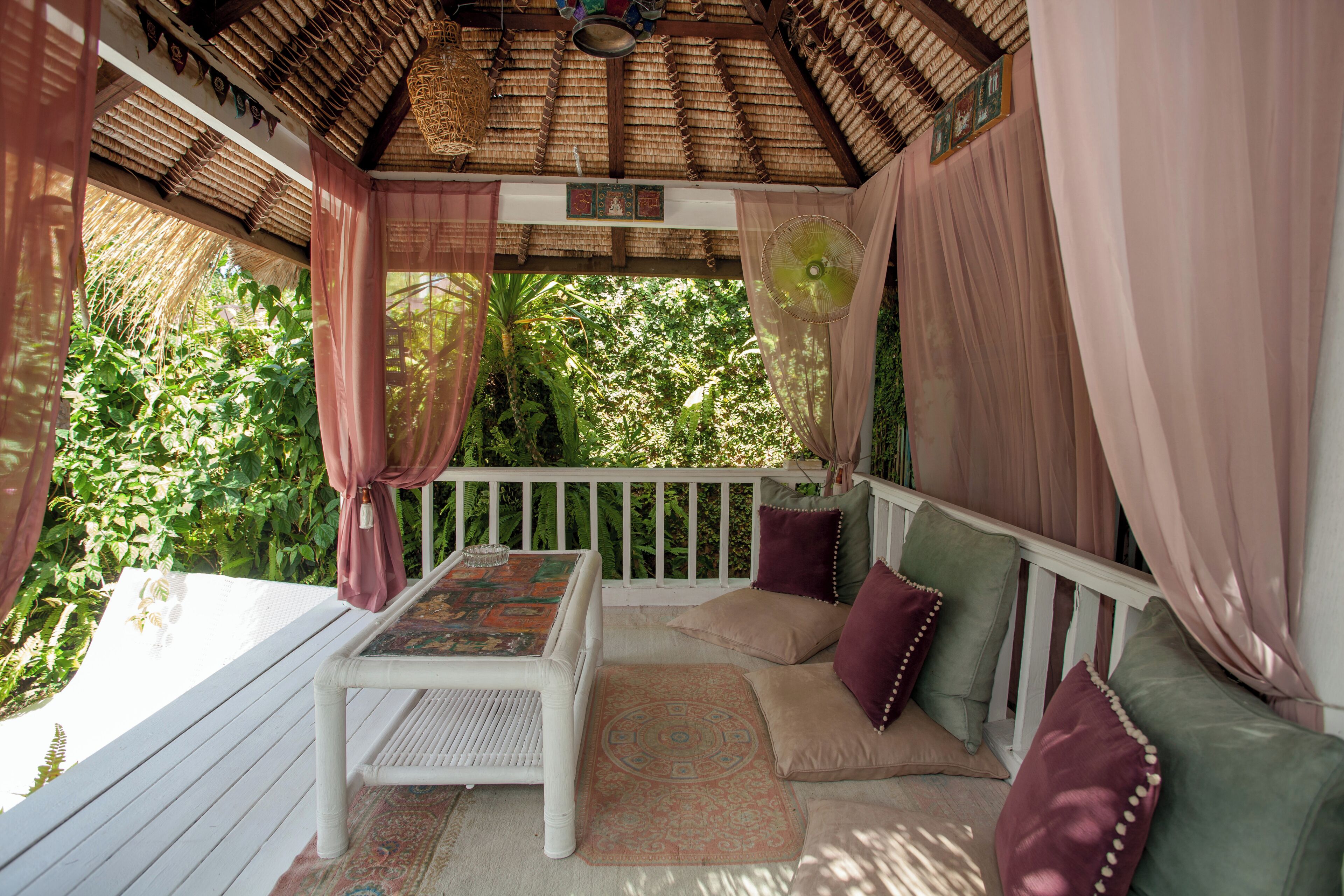A covered outdoor seating area with a table and cushions, surrounded by lush greenery.