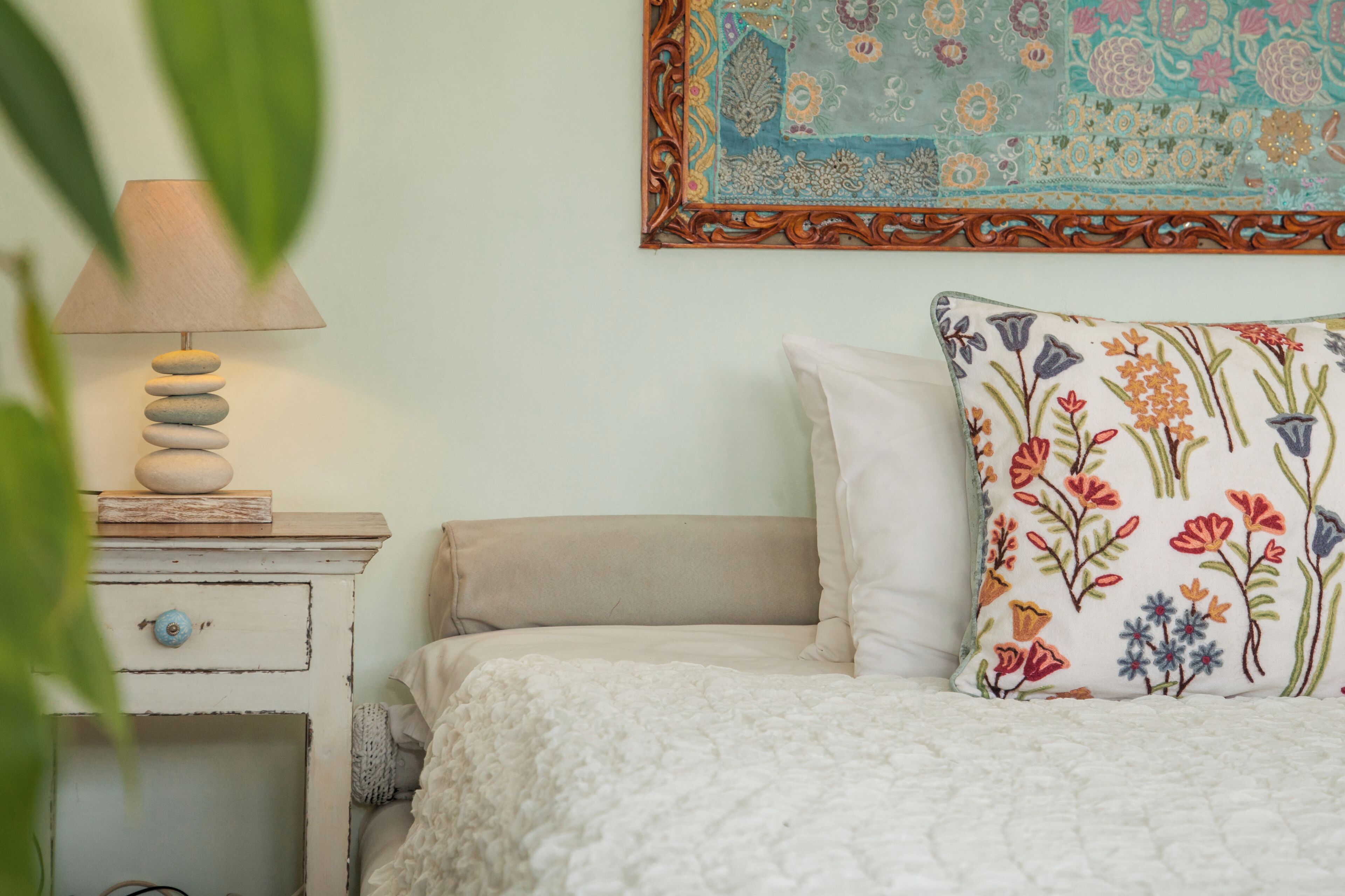 Una cama con un edredón blanco, un cojín decorativo con motivos florales, una lámpara de noche con base de madera y un tapiz con diseños florales.